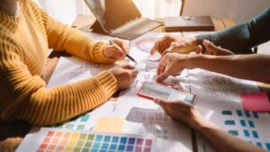 A team collaborating on design plans, with wireframe sketches, color swatches, and a tablet on a sunlit workspace.